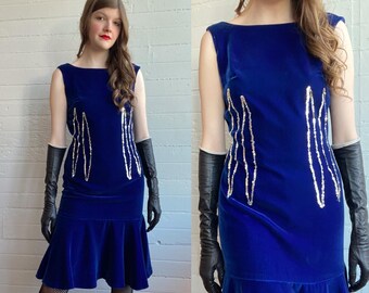 May include: A sleeveless, royal blue velvet dress with a flared hem. The dress features vertical silver sequin embellishments and is paired with long black gloves. The model is wearing black stockings and has red lipstick.