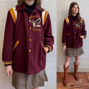May include: A maroon varsity jacket featuring yellow leather shoulder accents, yellow striped cuffs, and a front patch with an eagle graphic and the text Everett Eagles. The jacket has yellow snap buttons and two side pockets with yellow trim.