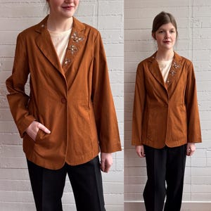 May include: A rust-colored blazer with a single-button closure and a notched lapel. The lapel is adorned with floral embellishments. The blazer is paired with black pants and a cream-colored top.