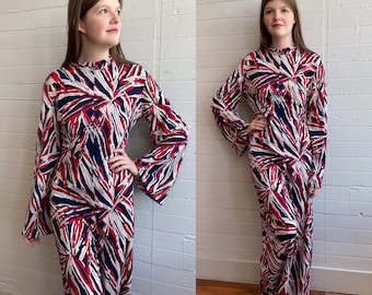 May include: A long-sleeved dress with a high neckline and a bold, abstract pattern in red, white, and navy blue. The dress has a flowing silhouette and wide sleeves, creating a striking visual effect. The fabric appears to be lightweight.