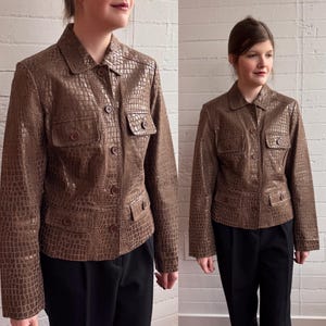 May include: A brown faux leather jacket with a crocodile skin effect. The jacket features a collared neckline, button closure, and four pockets. The jacket is worn with black trousers.
