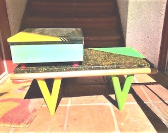 May include: A unique, colorful side table with a box-like top and a lower surface. The table features a teal box with a yellow and black top, a speckled surface, and V-shaped legs in yellow and green. The table is a retro design.