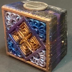 May include: A small, square, wooden box with a decorative design. The box is painted in shades of blue, purple, and gold. The design features intricate geometric patterns and a coin on top.