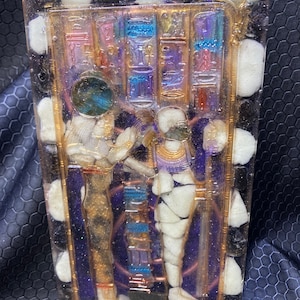 Könnte beinhalten: Ein Harz-Kunstwerk mit goldfarbenem und violettem Hintergrund. Das Stück zeigt zwei Figuren in Gold und Weiß, mit einem kreisförmigen Design in der Mitte. Die Figuren sind von weißen Steinen und einem violetten Hintergrund umgeben.