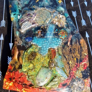May include: A resin art piece featuring a blue cross, gold leaf, and assorted stones and crystals. The piece is predominantly red, green, and blue.