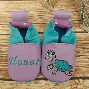 May include: A pair of baby shoes in lavender, teal, and purple. One shoe has the name "Hanae" embroidered in teal, and the other features a turtle design. The shoes have a pull tab and are made of soft material.