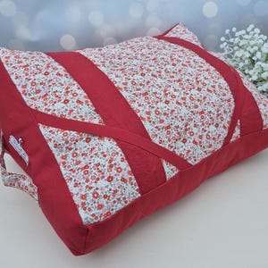 Peut inclure: Un coussin de sol rectangulaire composé d'un tissu à motif floral rouge et blanc contrasté par des panneaux et des sangles en tissu rouge uni. Le coussin présente une poignée latérale et une petite étiquette blanche avec du texte rouge et bleu.