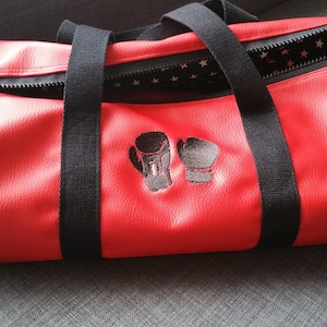 Peut inclure: Sac de sport en cuir rouge avec des sangles noires et une fermeture à glissière. Le sac est orné d'un motif brodé noir de deux paires de gants de boxe.