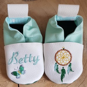 May include: A pair of light blue baby shoes with white leather soles. One shoe is embroidered with the name "Betty" and a butterfly. The other shoe is embroidered with a dreamcatcher.
