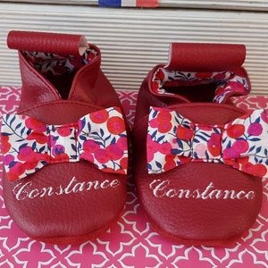 Puede incluir: Un par de zapatos de bebé de cuero rojo con un lazo de estampado floral y el nombre "Constance" bordado en cada zapato.