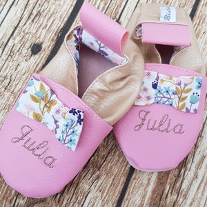 Puede incluir: Zapatos de bebé de cuero rosa con un lazo floral y el nombre "Julia" bordado en la parte delantera.