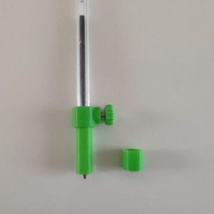 Puede incluir: Un bolígrafo de plástico verde con un tubo de plástico transparente y un cartucho de tinta negro. El bolígrafo tiene un pequeño botón en el lateral y un tapón de plástico verde separado.