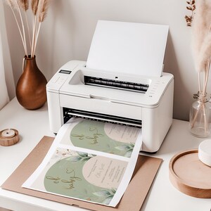 Op de afbeelding: Een witte desktop-printer die een vel papier uitwerpt met twee geprinte baby shower uitnodigingen. De uitnodigingen hebben een saliegroen boogontwerp, goudkleurige cursieve tekst met oh baby en botanische illustraties van groene bladeren.