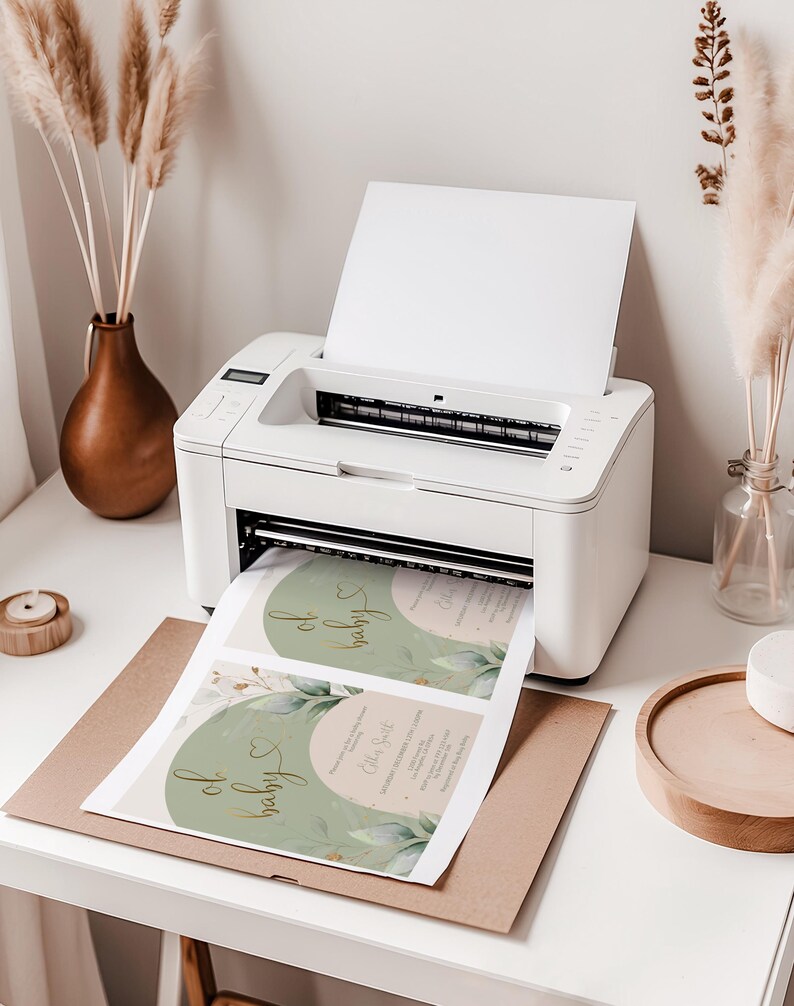 Op de afbeelding: Een witte desktop-printer die een vel papier uitwerpt met twee geprinte baby shower uitnodigingen. De uitnodigingen hebben een saliegroen boogontwerp, goudkleurige cursieve tekst met oh baby en botanische illustraties van groene bladeren.