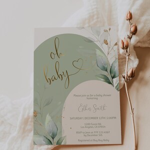 Op de afbeelding: Een rechthoekige uitnodigingskaart voor een babyshower met een saliegroen boogontwerp, goudfolie tekst met oh baby en aquarel eucalyptusbladeren. De kaart bevat de evenementgegevens voor Esther Smith op 12 december om 14:00 uur.