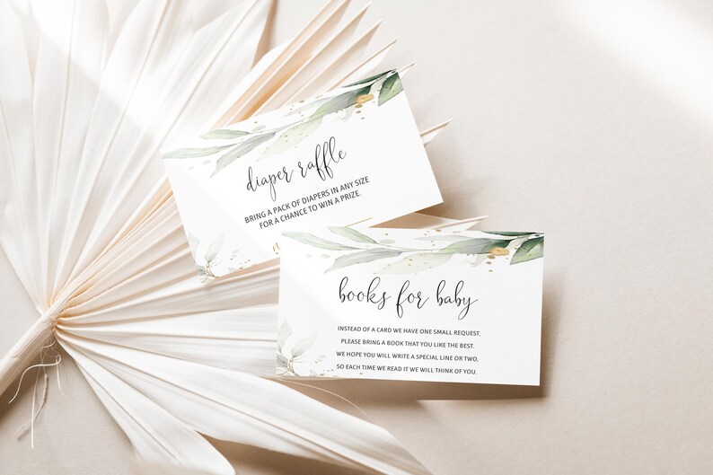 May include: Two white baby shower cards with elegant script, one reading "diaper raffle" and the other "books for baby." Both cards feature watercolor greenery and gold accents. The cards are set against a neutral, textured background.