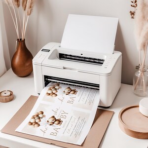 May include: A white printer printing baby shower invitations. The invitations feature a teddy bear holding balloons and the text "He or She, What Will Baby Be?" on a light beige background. The printer sits on a white desk.