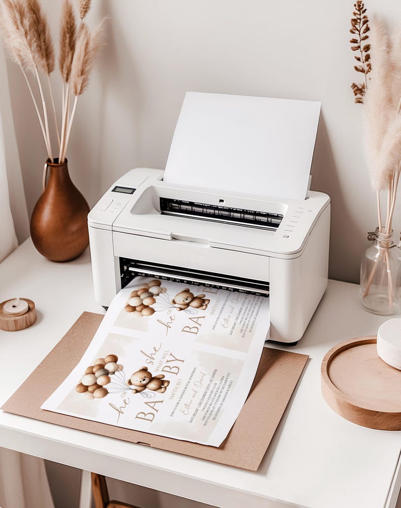 May include: A white printer printing baby shower invitations. The invitations feature a teddy bear holding balloons and the text "He or She, What Will Baby Be?" on a light beige background. The printer sits on a white desk.