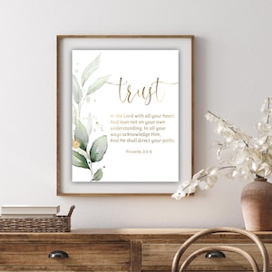 Può includere: Una stampa incorniciata con la parola "trust" in oro, accompagnata da un versetto biblico e vegetazione ad acquerello. L'opera è esposta su una cassettiera in legno, con un vaso di fiori bianchi a destra.