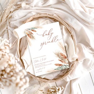 K&ouml;nnte beinhalten: Eine Baby-Sprinkle-Einladungskarte mit den Worten "baby sprinkle" in eleganter brauner Schrift. Die Karte zeigt florale und Pampasgras-Akzente in Braun-, Beige- und Gr&uuml;nt&ouml;nen. Die Einladung befindet sich in einem geflochtenen Korb.