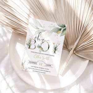 May include: A baby shower invitation with the words "oh BOY" in gold and green, surrounded by watercolor leaves and gold accents. The invitation is on a white plate with a dried palm leaf in the background.