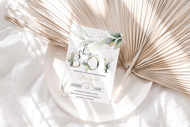 May include: A baby shower invitation with the words "oh BOY" in gold and green, surrounded by watercolor leaves and gold accents. The invitation is on a white plate with a dried palm leaf in the background.