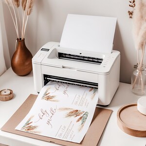 K&ouml;nnte beinhalten: Ein wei&szlig;er Drucker druckt Baby-Sprinkle-Einladungen. Die Einladungen haben ein Blumendesign mit den Worten "baby sprinkle" in einer Schreibschrift. Der Drucker steht auf einem wei&szlig;en Schreibtisch mit dekorativen Vasen und einem Holztablett.