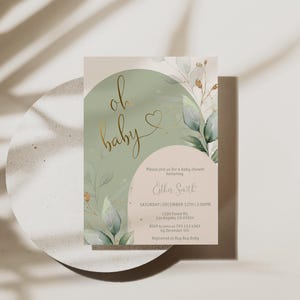 Op de afbeelding: Een rechthoekige uitnodigingskaart voor een babyshower met een saliegroen boogontwerp, goudfolie tekst oh baby en botanische aquarelillustraties. De kaart bevat gedrukte evenementdetails voor Esther Smith op een lichtbeige achtergrond.