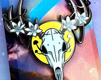 Può includere: Spilla smaltata raffigurante un teschio di cervo con corna, ornata da fiori bianchi e funghi, su un cerchio giallo. La spilla fa parte di "A Nature Pin Series" e la parola "Rebirth" è visibile.