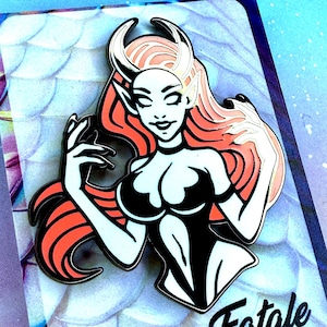 May include: A black and white enamel pin featuring a stylized woman with red hair, horns, and a black outfit. The pin has a Femme Fatale theme, with the text "Femme Fatale" at the bottom.