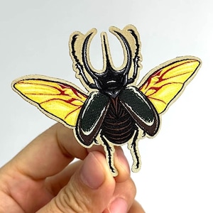 May include: Embroidered patch featuring a detailed illustration of a beetle with yellow and red-veined wings. The beetle has a black body, green accents, and a tan border. The patch is held by a hand. Text: Art by: JBlake Design Instagram @jbzydraws.