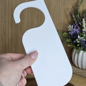 May include: A white door hanger with a curved top and a rectangular body. The door hanger is held by a hand. The background features a wooden surface and a vase with flowers.