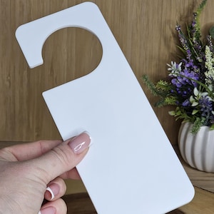 May include: A white door hanger with a curved top and a large circular cutout. The rectangular body is held by a hand with manicured nails. The background is a wooden surface with a vase of flowers.
