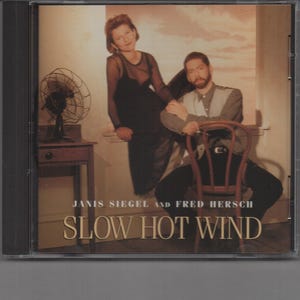 May include: A CD cover featuring a black and white photo of two people, a woman in a black dress and a man in a light-colored shirt, sitting on a chair. The text on the cover reads "Janis Siegel and Fred Hersch Slow Hot Wind".