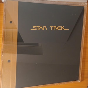 May include: A black and gold Star Trek binder in a clear plastic case. The words "STAR TREK" are printed in gold on the black cover. The binder has a gold spine and is on a wooden surface.
