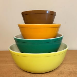 May include: A set of four nested mixing bowls stacked vertically. The bowls feature a smooth, solid-colored exterior in descending order of size from top to bottom: dark brown, bright orange, forest green, and a vibrant lemon yellow.