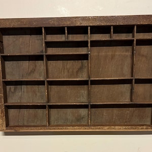 May include: A vintage wooden printer's tray with multiple small compartments. The tray is rectangular with a dark brown finish and a grid-like interior. The wood shows signs of wear, adding to its antique charm.