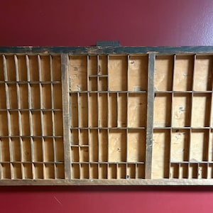 May include: A rectangular wooden printer's type case featuring a grid of numerous small, open compartments of varying sizes. The frame is dark brown with a weathered finish, while the interior dividers are a lighter, natural wood tone.