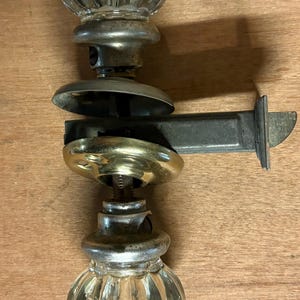 May include: Antique door hardware set featuring two clear, faceted glass knobs with metal bases and a connecting metal bar. The hardware has a vintage aesthetic, with signs of wear and age.