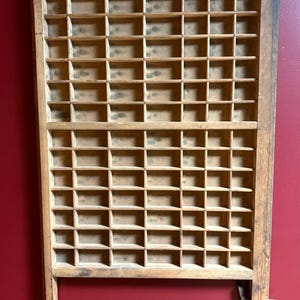 Könnte beinhalten: Ein Vintage-Druckertablett aus Holz mit zahlreichen kleinen Fächern. Das Tablett besteht aus hellem Holz mit einer dunklen Oberseite und einem dunklen Rahmen. Es ist für kleine Gegenstände oder Buchstaben konzipiert und an einer roten Wand befestigt.