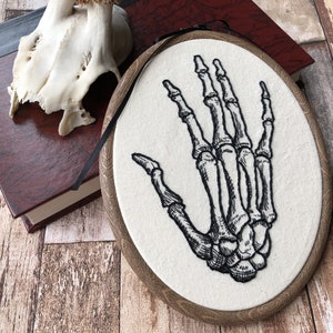 An oval embroidery hoop. A detailed embroidery of the bones of the hand is on the ivory fabric. The hoop is leaning against a book. A deer skull is on the book.