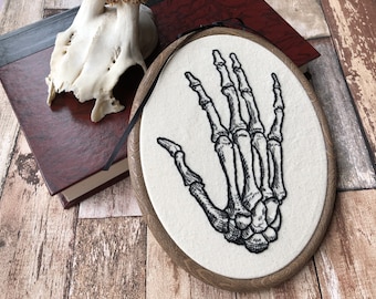 May include: A black and white embroidered skeletal hand on a white fabric background. The embroidery is in a wooden hoop with a dark brown wood finish.