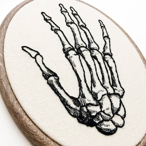 An oval embroidery hoop. A detailed embroidery of the bones of the hand is on the ivory fabric.