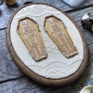 May include: Two embroidered skeletons in gold and white are inside oval-shaped coffins, stitched onto a white fabric background with a lace border. The fabric is framed in a dark wood frame.