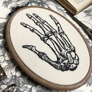 An oval embroidery hoop. A detailed embroidery of the bones of the hand is on the ivory fabric.