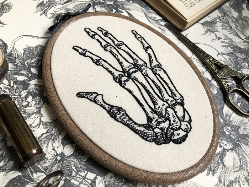 An oval embroidery hoop. A detailed embroidery of the bones of the hand is on the ivory fabric.