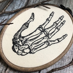 An oval embroidery hoop. A detailed embroidery of the bones of the hand is on the ivory fabric.