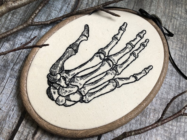 An oval embroidery hoop. A detailed embroidery of the bones of the hand is on the ivory fabric.