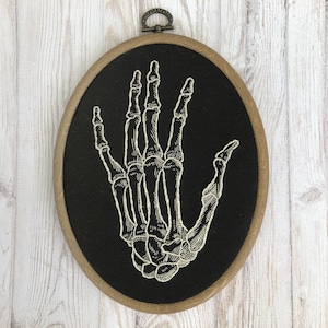 May include: Embroidered skeletal hand on black fabric in a wooden embroidery hoop.