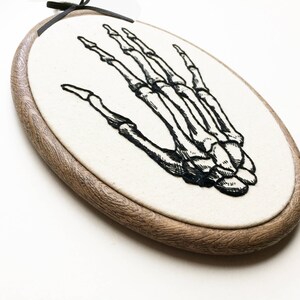 An oval embroidery hoop. A detailed embroidery of the bones of the hand is on the ivory fabric.